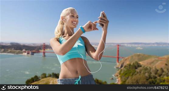 sport, technology and healthy lifestyle concept - smiling young woman with smartphone, earphones and fitness tracker listening to music over golden gate bridge in san francisco bay background. woman with smartphone and earphones doing sports. woman with smartphone and earphones doing sports
