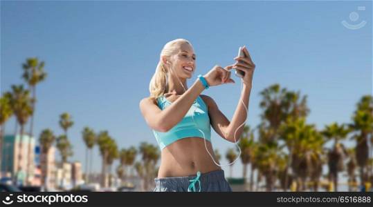 sport, technology and healthy lifestyle concept - smiling young woman with smartphone, earphones and fitness tracker listening to music and exercising over venice beach background in california. woman with smartphone and earphones doing sports. woman with smartphone and earphones doing sports