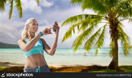 sport, technology and healthy lifestyle concept - smiling young woman with smartphone and fitness tracker listening to music and exercising over tropical beach background in french polynesia. woman with smartphone and earphones doing sports. woman with smartphone and earphones doing sports