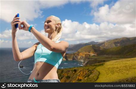 sport, technology and healthy lifestyle concept - smiling young woman with smartphone, earphones and fitness tracker listening to music over big sur hills and pacific ocean background in california. happy woman with smartphone and earphones outdoors. happy woman with smartphone and earphones outdoors