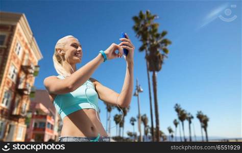 sport, technology and healthy lifestyle concept - smiling young woman with smartphone, earphones and fitness tracker listening to music over venice beach background in california. happy woman with smartphone and earphones outdoors. happy woman with smartphone and earphones outdoors