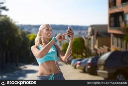 sport, technology and healthy lifestyle concept - smiling young woman with smartphone, earphones and fitness tracker listening to music and exercising over san francisco city background. woman with smartphone and earphones doing sports. woman with smartphone and earphones doing sports