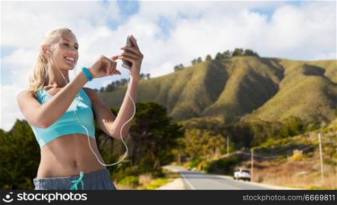 sport, technology and healthy lifestyle concept - smiling young woman with smartphone, earphones and fitness tracker listening to music over big sur hills and road background in california. woman with smartphone and earphones doing sports. woman with smartphone and earphones doing sports