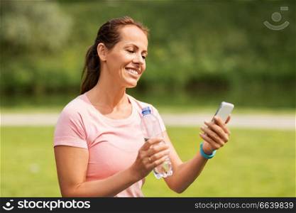 sport, technology and healthy lifestyle concept - woman with smartphone and fitness tracker drinking water after exercising in park. woman with smartphone drinking water in park