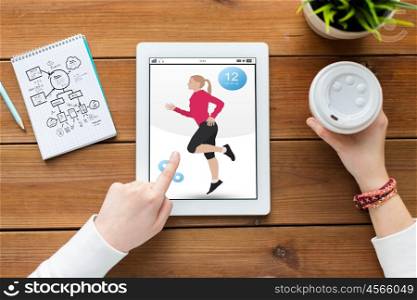 sport, technology and people concept - close up of woman with fitness application on tablet pc computer screen, notebook and coffee on wooden table