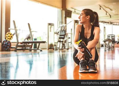 Sport woman relax resting after workout or exercise in fitness gym. Sitting and drinking protein shake or drinking water on floor. Strength training and Bodybuilder muscle theme. Warm and cool tone