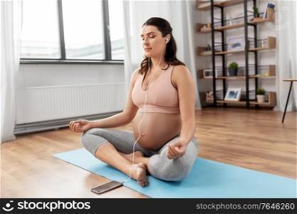 sport, yoga and people concept - happy pregnant woman with earphones and smartphone listening to music and meditating at home. pregnant woman with earphones doing yoga at home