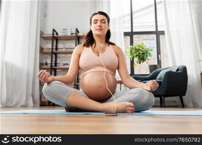 sport, yoga and people concept - happy pregnant woman with earphones and smartphone listening to music and meditating at home. pregnant woman with earphones doing yoga at home
