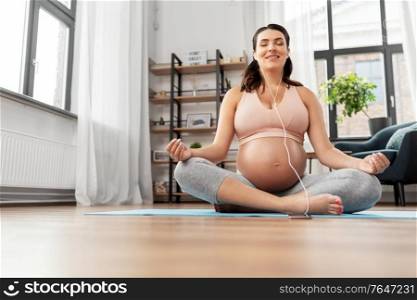 sport, yoga and people concept - happy smiling pregnant woman with earphones and smartphone listening to music and meditating at home. pregnant woman with earphones doing yoga at home