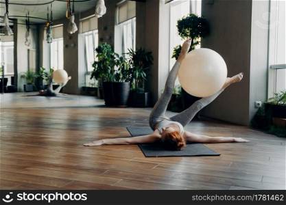 Sportive female lying on yoga mat with outstretched arms holding exercise ball between legs while exercising in modern fitness studio with big mirror, doing stability ball exercises. Pilates concept. Fit woman training with exercise ball on yoga mat in fitness studio