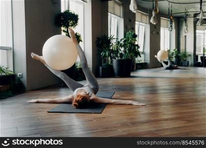 Sportive female lying on yoga mat with outstretched arms holding exercise ball between legs while exercising in modern fitness studio with big mirror, doing stability ball exercises. Pilates concept. Fit woman training with exercise ball on yoga mat in fitness studio
