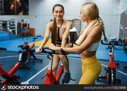 Sportive girlfriends doing exercise on stationary bikes in gym, front view. People on fitness workout in sport club, athletic girls in sportswear on training indoors. Sportive girlfriends on stationary bikes in gym