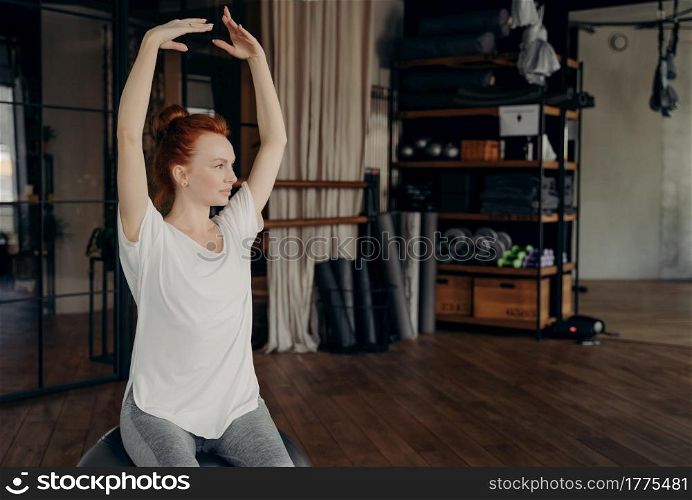 Sportive gracile young woman with red hair sits on big silver pilates ball with hands lifted over head warming up before exercising in fitness studio. Concept of healthy lifestyle and sport indoor. Sportive young woman with red hair sits on big pilates ball with hands lifted over head