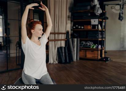 Sportive gracile young woman with red hair sits on big silver pilates ball with hands lifted over head warming up before exercising in fitness studio. Concept of healthy lifestyle and sport indoor. Sportive young woman with red hair sits on big pilates ball with hands lifted over head