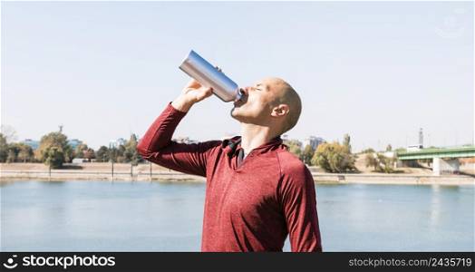 sportive young man drinking water from bottle standing near lake