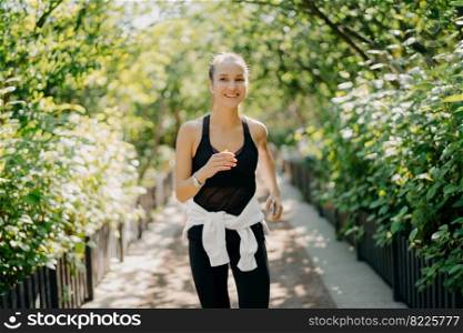 Sportive young woman has jogging workout training runs outdoor smiles pleasantly enjoys good day dressed in sportsclothes listens music from playlist. Start day from morning jog. Female runner
