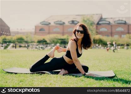 Sports activity concept. Horizontal shot of happy motivated woman stretches on fitness mat trains outdoors during summer day has sporty healthy body wears sunglasses cropped top and leggings