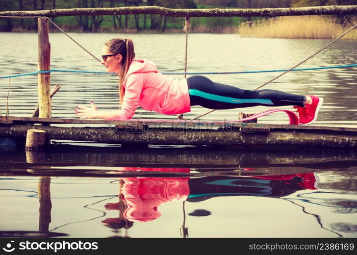 Sports and activities concept. Slim fit attractive woman exercising stretching outdoor. Young motivated girl training in sporty clothes on lake shore. Girl training in sporty clothes on lake shore