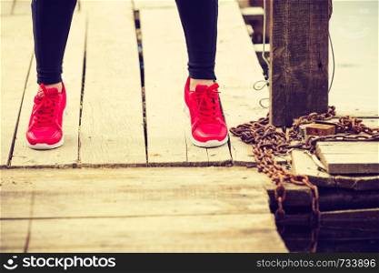 Sports and activities. Part body of training active sporty girl outside. Slim fit legs wearing red shoes on wooden pier, lake shore. Slim fit legs wearing red sport shoes on wooden pier
