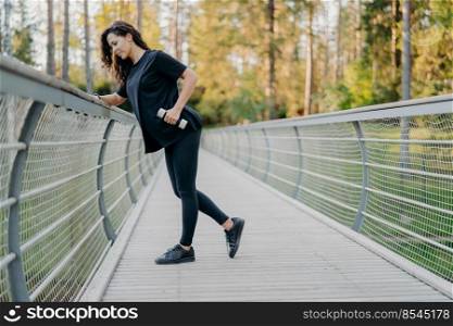 Sportswoman holds dumbbells and does workout outdoor, dressed in active wear, poses on bridge and looks down, forest in background. Sporty healthy strong female trains muscles during exercising