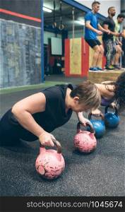 Sportswomen doing push-ups with kettlebells with mates doing box jumps in background. Sportswomen doing push-ups with kettlebells