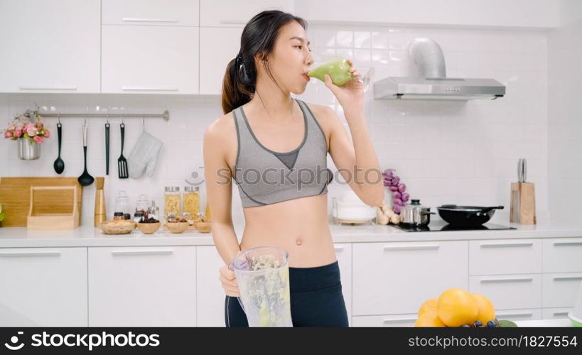 Sporty Asian woman drinking apple juice in the kitchen, beautiful female in sport clothing use organic fruits lots of nutrition making apple juice by herself at home. Healthy food concept.