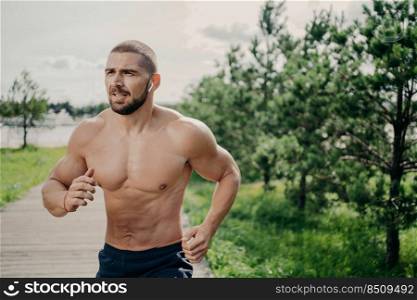 Sporty athletic man runs outdoor with naked torso, leads healthy lifestyle, has morning workout, listens music in wireless earphones, has muscular body, breathes fresh air. Health activities