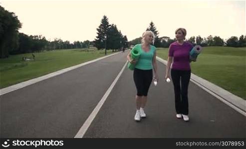 Sporty attractive adult female friends in sport clothes holding exercise mats and bottles of water walking through the park for a workout. Beautiful women going to exercise outdoors. Slow motion. Stedicam stabilized shot.