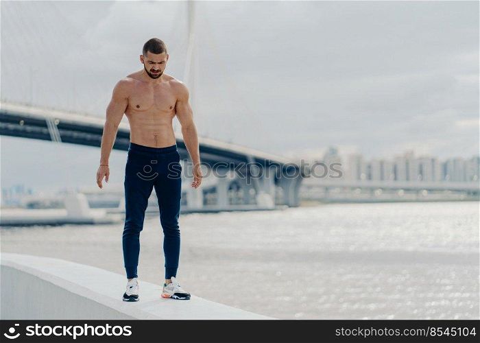 Sporty bearded man with muscular body walks near river, dressed in sport trousers and sneakers, looks down, has muscular body, stands against beautiful landscape. Athlete bodybuilder poses outside