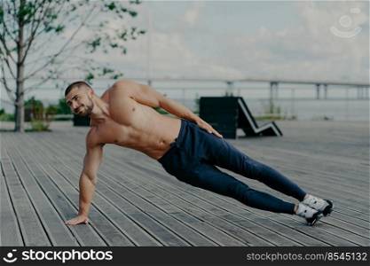 Sporty bodybuilder stands in side plank, exercises outdoor and wears sport clothes, leads healthy lifestyle, has fit body and muscles. Outdoor shot of determined sportsman does health activities
