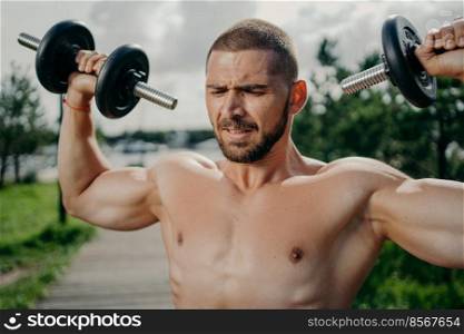 Sporty determined muscular European man exercsises with dumbbells, makes weightlifting outdoor, has naked torso, trains muscles, wants to have athletic body. Strength and motivation concept.