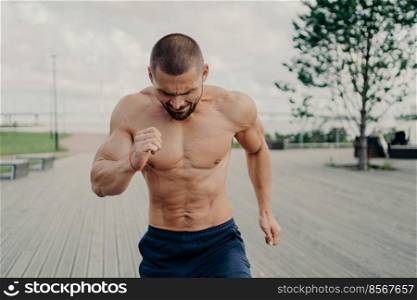 Sporty energized bearded male runner goes jogging on walkway next to river or sea, has daily training, poses with naked torso, puts all efforts in staying fit and healthy. Morning workout concept