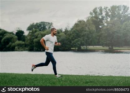 Sporty European man dressed in t shirt, trousers and sneakers and runs along river, trains outdoors, leads active lifestyle, breathes fresh air. People, sport, wellness and recreation concept.