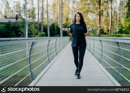 Sporty European woman in black t shirt and leggings, walks across bridge, holds two dumbbells, works on training biceps, trains outdoor, has thoughtful expression, poses with sport equipment