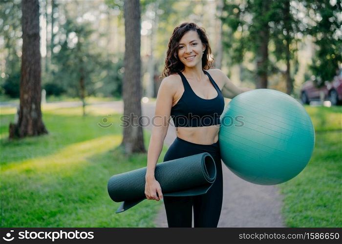 Sporty European woman in cropped top and leggings, carries rolled up karemat and fitness ball, going to have aerobics exercises, poses outdoor against green nature background, has pilattes class