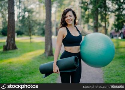 Sporty European woman in cropped top and leggings, carries rolled up karemat and fitness ball, going to have aerobics exercises, poses outdoor against green nature background, has pilattes class