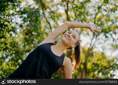 Sporty fitness woman does warm up and stretching exercises leans aside has content face expression dressed in active wear poses against nature background during summer day. Sport and motivation