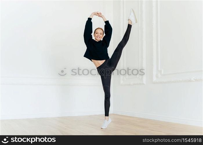 Sporty flexible female ballerina raises hands, balances on one leg, has cheerful facial expression, being in good body shape, demonstrates yoga pose, wears black hoody, leggings and sportshoes