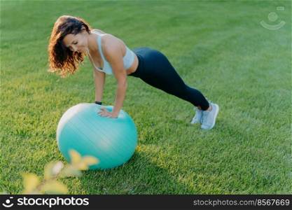 Sporty girl stands in plank pose on gymnastic ball, dressed in sportswear, does physical exercises on green lawn outdoor, keeps fit and healthy. People, yoga, flexibility, lifestyle concept.