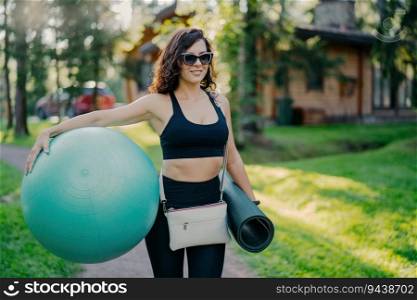 Sporty happy brunette holds fitness ball and karemat, dressed in cropped top and leggings. Preparing for outdoor fitness exercises by a private house, on green grass.