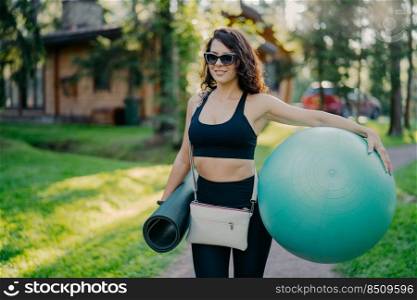 Sporty happy curly brunette woman wears cropped top and leggings holds fitness ball and rolled up karemat, going to have fitness exercises in open air poses against private house and green grass