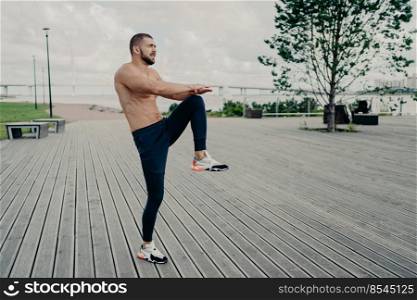 Sporty man does sport exercises outdoors in morning, raises legs, wears active wear, warms up before cardio training. Successful sportsman exercises in open air. Physically active man has workout