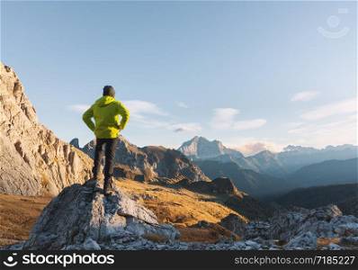 Sporty man standing on the stone against mountain valley at sunset in spring. Happy young man, rocks, forest and blue sky in Dolomites, Italy. Traveler hiking in alpine mountains. Travel and tourism