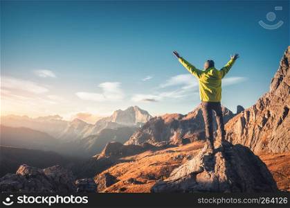 Sporty man standing on the stone with raised up arms against mountain valley at sunset. Happy young man, rocks, autumn forest and blue sky in Dolomites, Italy. Traveler hiking in alpine mountains