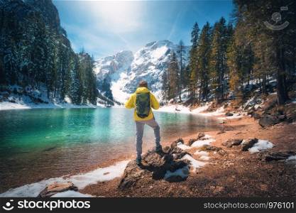 Sporty man with backpack is standing on the stone near lake with azure water at sunny day in autumn. Travel. Landscape with guy, reflection in water, snowy mountains, orange trees, blue sky in fall