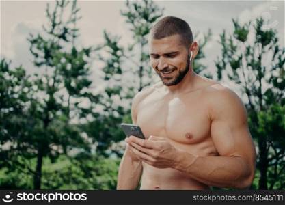Sporty man with strong muscular body, uses smartphones and wireless earphones, downloads music for training, has thick bristle, poses outdoor against nature background. Sportsman uses modern gadget