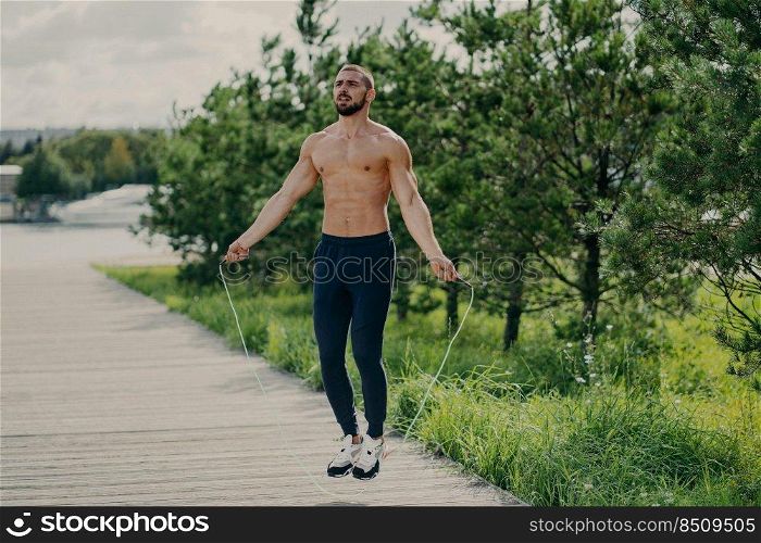 Sporty man with thick beard does exercises with jumping rope, has muscular torso, stays in good physical shape, poses outdoor. Fitness and healthy concept. Male runner warms up with sport equipment
