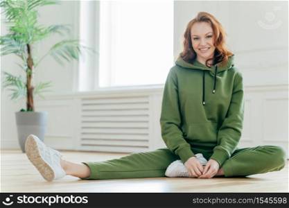 Sporty motivated female demonstrates nice flexibility, slim body, stretches legs, has fitness pilates exercises, wears green tracksuit, white sneakes, sits on floor, prefers healthy lifestyle.
