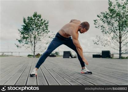 Sporty motivated handsome man leans and does sport exercises, poses outdoor and busy doing workout. Determined shirtless guy wears sneakers, has morning training in park. Healthy lifestyle concept