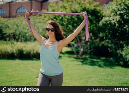 Sporty pretty woman in sportsclothes does exercise workout with elastic rubber bandage gum wears sunglasses poses outdoor against green background. People healthy lifestyle stretching before sport